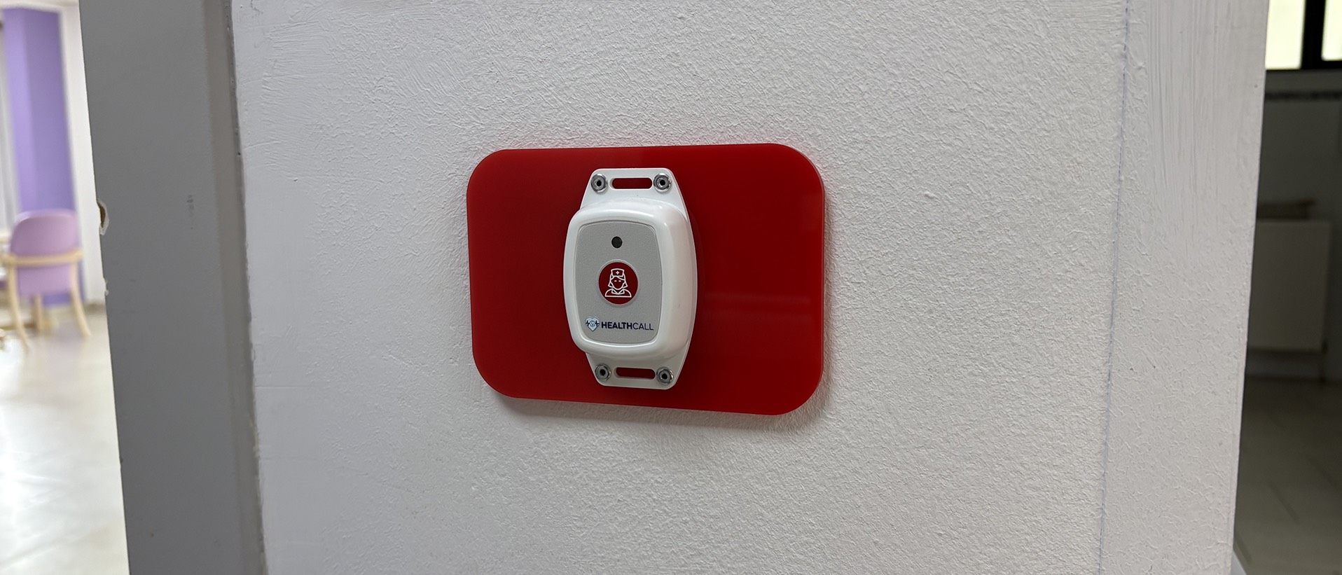 Draadloze Bluetooth-oproepknop Healthcall bevestigd op een rode acrylafwerkingsplaat die de plek van de oude Televic-knop afdekt, op een witte gangmuur