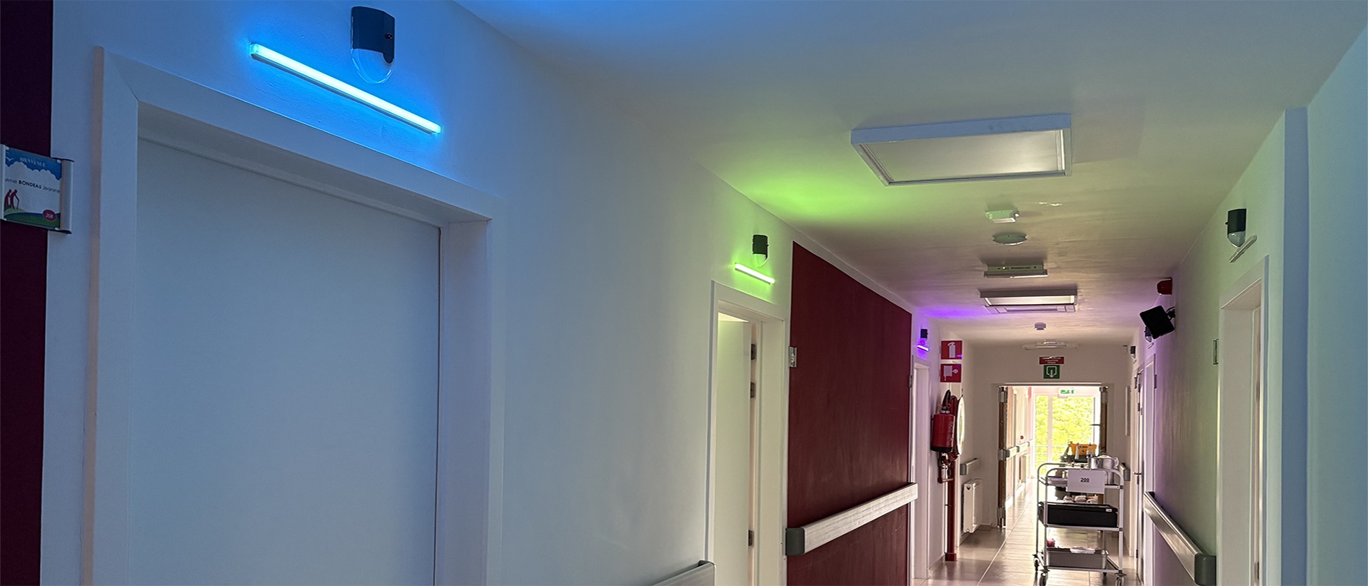 Verdiepingsgang van woonzorgcentrum Le Petit Paradis bij nacht gezien, LED-paneeltjes van 50 cm boven elke kamerdeur in cyaan, groen en magenta, kalme nachtsfeer