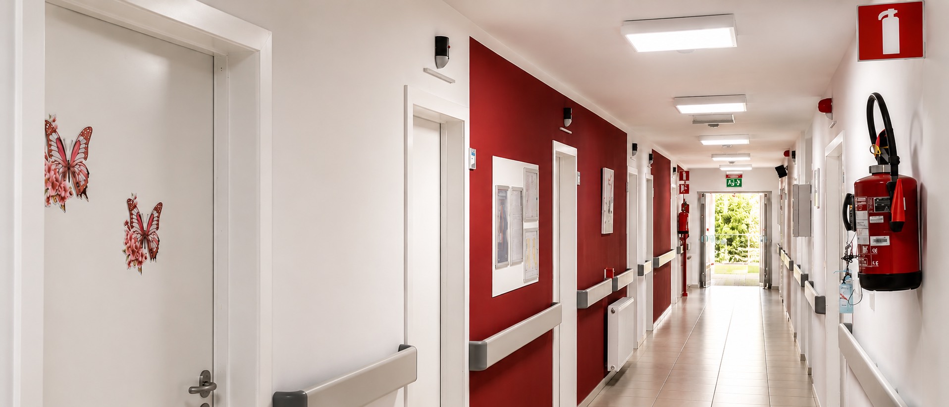 Verdiepingsgang van woonzorgcentrum Le Petit Paradis, witte muren met rode accenten, Healthcall LED-paneeltjes aan het plafond, natuurlijk licht aan het einde van de gang