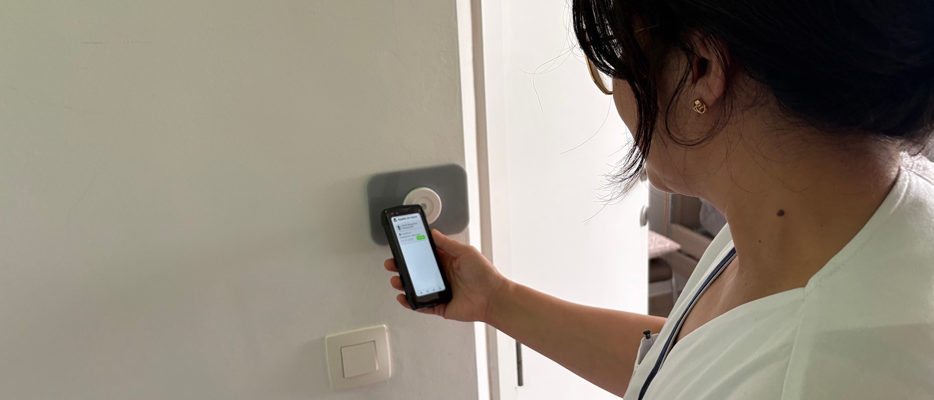 Zorgverleenster in lichte kiel voor de ingang van een kamer, industriële smartphone van 4,5 inch in de hand met de Healthcall-app, naast de NFC-lezer aan de muur