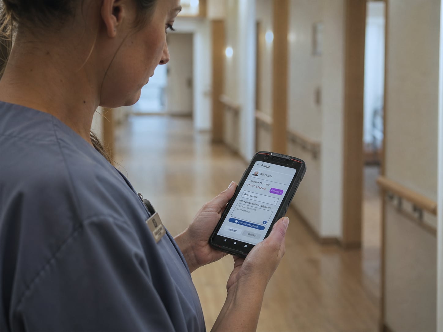 Care worker in navy polo consulting a nurse-call alert on a shared professional Android smartphone per duty station, corridor of a Belgian care home equipped with Healthcall